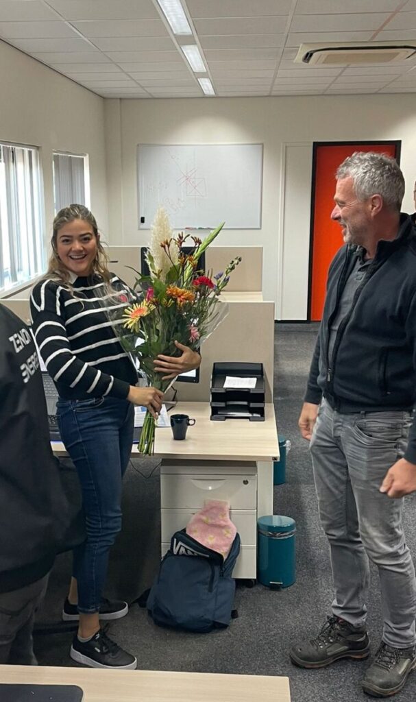 Marcel and Veronica stand at the desk with flowers in veronicas hands 